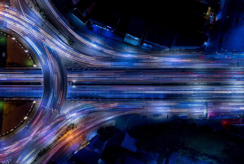 Aerial view above a multi lane highway with a roundabout. It is dark so street lights and lights from cars make out the road. Purple hues and dark surroundings. 